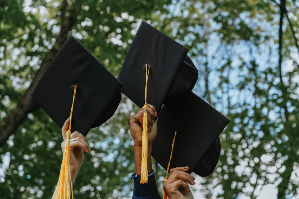 Three people with different skin tones hold graduation caps aloft with tree branches in the background.