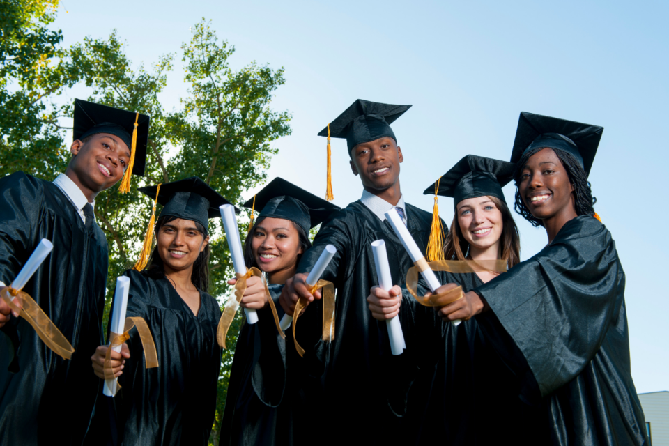 Image shows six students in graduation regalia. There are 2 men and four women with different skin tones. All are holding their rolled up diploma out in front of them towards the camera and smiling.